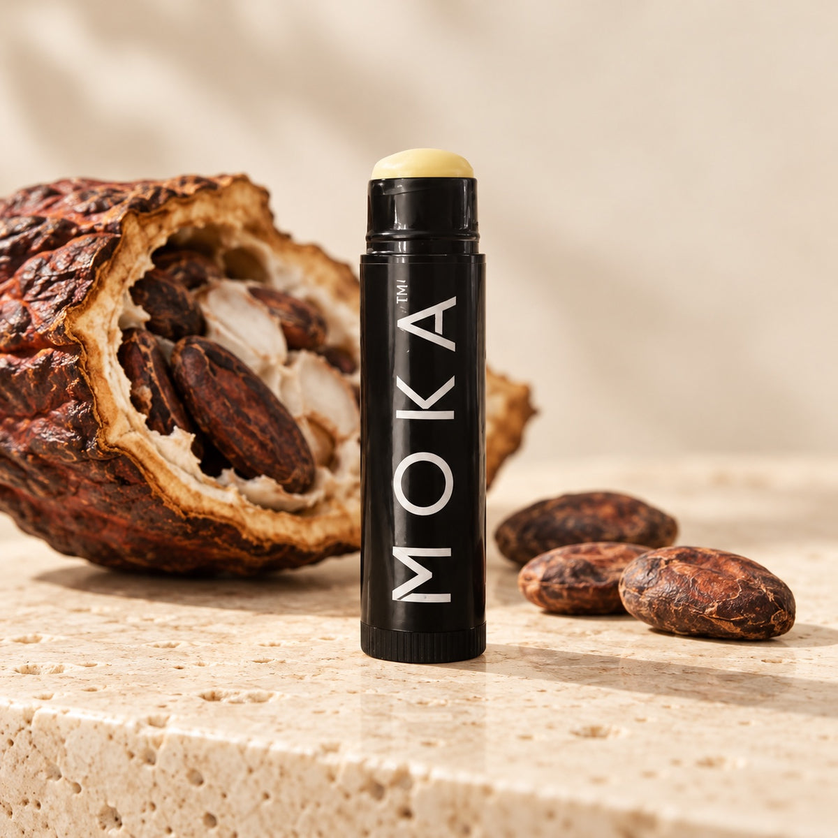 MOKA lip balm standing on travertine beside a cacao pod and cacao beans