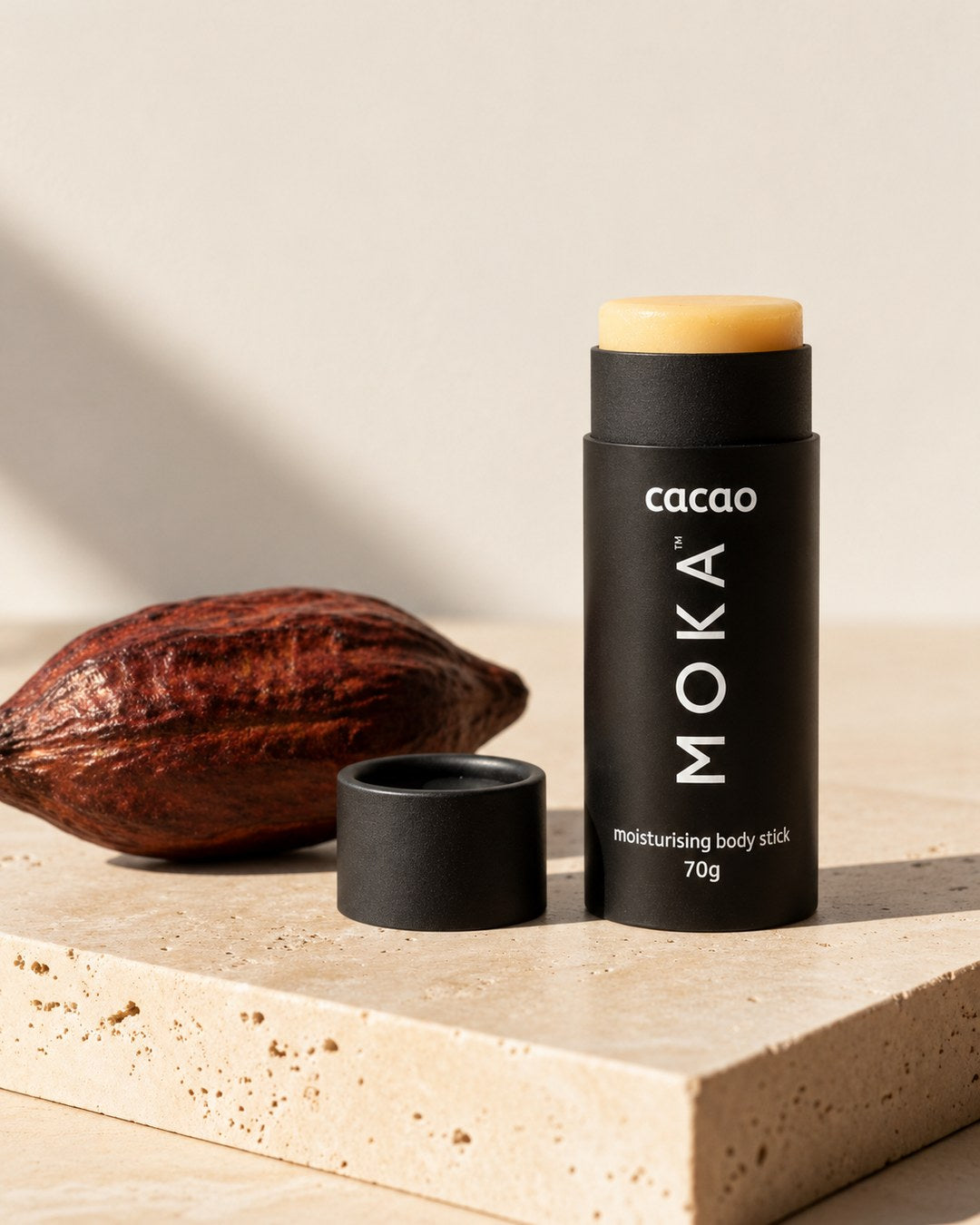 The MOKA cacao body butter stick, lid off, beside a cacao pod on travertine stone